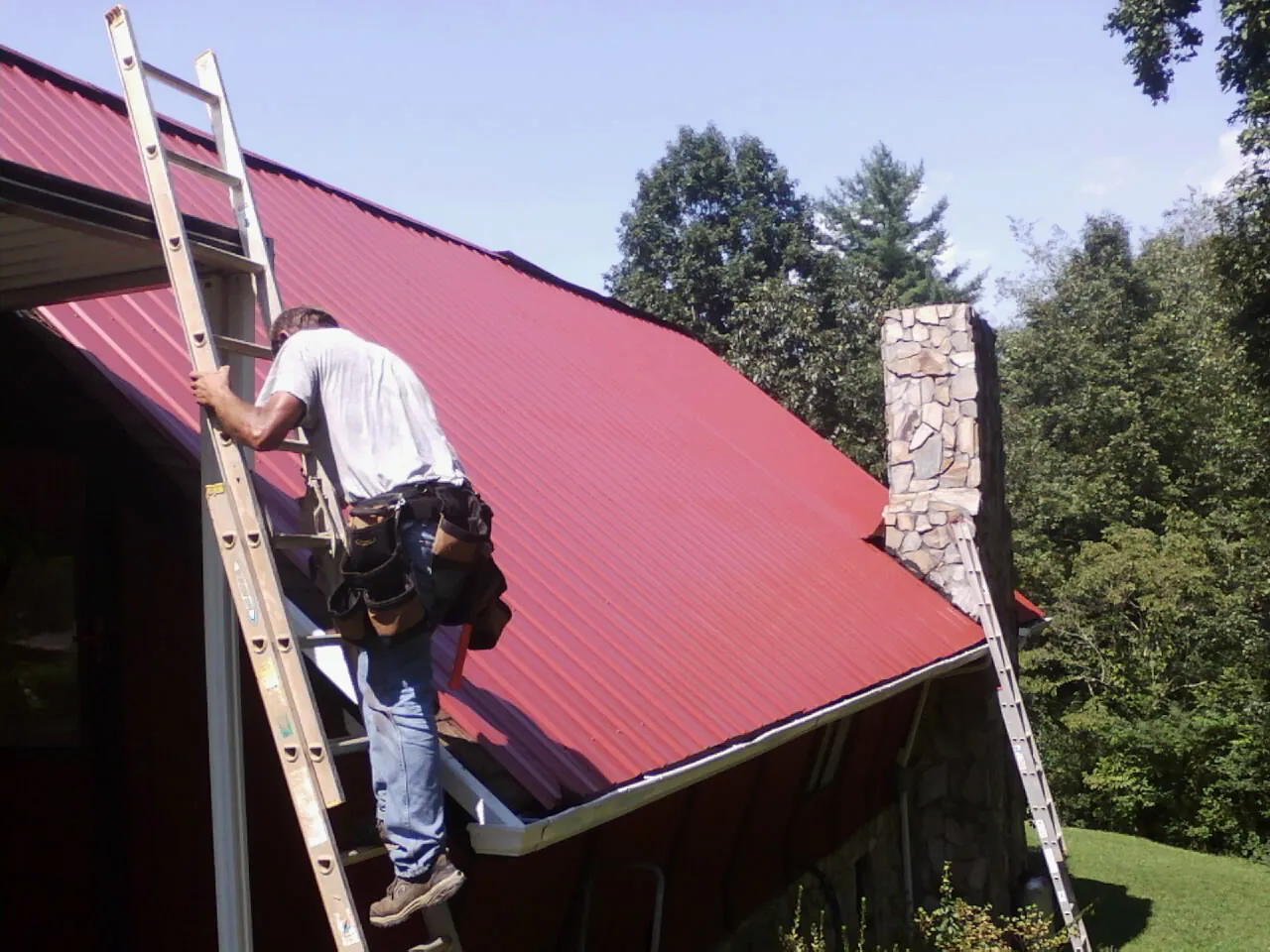 Roofer on a ladder beside a red metal roof and stone chimney — Soffit & Fascia Repair by Roofing Brothers in Carpenter